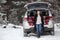 Adult woman in mink fur coat sitting in trunk of car under opened back door, wintry forest