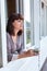 Adult woman in the morning holding a cup of tea or coffee and looking away standing near the window in her home