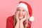 An adult woman in a Santa hat rests her chin with her hands on a pink background, close-up