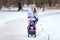 Adult woman walking with her baby on winter park, pulling sled with daughter on snow pathway, copyspace