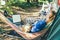 Adult woman working on laptop pc while lying in hammock, with her cockapoo puppy near motorhome on camping trip. Female