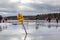 Adults and children playing ice hockey on lake, warning sign in the foreground.