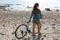 Adventure on bike. Back view portrait of young woman walking with bicycle on wild beach enjoying view of Atlantic Ocean