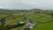 Aerial ascending shot of group of vehicles driving on road in village in countryside. Green grasslands divided by rows