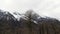 Aerial of bald tree tops and high snowy mountains covered by heavy clouds. Stock footage. Winter landscape with high