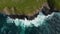 Aerial birds eye overhead top down ascending view of rough waves crashing on rocky coast. Green grass on top of cliffs