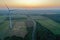 Aerial drone perspective view on wind turbine, snake shape road, wheat fields and forest during summer sunset