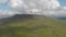 An aerial forward footage of a rocky summit mountain with green slope and trail path under a majestic blue sky