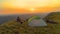 AERIAL: Golden sun rays shine on puppy and couple cuddling by tent at sunset.