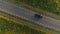 Aerial shot from above driving black car in a field along a rural road in summer at sunset.