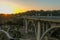 Aerial shot of the Colorado Street Bridge surrounded by homes, lush green trees, grass and plants, majestic mountains at sunset