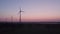 Aerial shot by a drone of windmills with rotating wings among green fields against a sunrise or sunset background. Wind