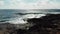 Aerial shot over sea waves hitting rocky beach splashing against rocks. Blue sky with sun road on the horizon. Ocean on a windy da