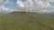 An aerial sideway left to right footage of a rocky summit mountain with green slope and trail path under a majestic blue sky