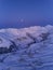 Aerial of snowy mountain range on winter sunrise at ski resort. Moon above mountains valley and village with switchbacks