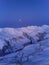 Aerial of snowy mountain range on winter sunrise at ski resort. Moon above mountains valley and village with switchbacks