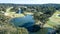 Aerial view of golf course fairways and green with flag, bunkers and dam water hazard surrounded by trees in background