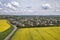Aerial view of ground road in green fields with blooming rapeseed plants, suburb houses on horizon and blue sky copy space