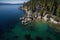 an aerial view of a lake surrounded by trees and rocks with clear blue water in the foreground and a forested area in the