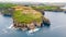 Aerial view of a large rocky ledge of the island in the ocean with green and yellow fields and a small lighthouse