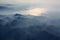 Aerial view of misty mountains, lake and clouds above the mountain peaks, opposite the sunlight, blue tinted