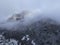 Aerial view of mountain with boundary tower, forest underneath fog, slopes with snow covered in Switzerland, cloudy