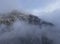 Aerial view of mountain with boundary tower, forest underneath fog, slopes with snow covered in Switzerland, cloudy