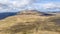 An aerial view of a Scottish mountain summit plateau with heather, trail path and huge cliff under a majestic blue sky and huge