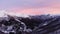 Aerial view of snow covered mountain range during pink and yellow sunset or sunrise. Dark forest in the valley. slow