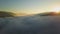 Aerial view of vibrant sunset over white dense foggy clouds with distant dark silhouettes of mountains on horizon.