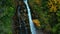 Aerial view of waterfall in mixed forest in the mountains. Video from drone flying over spruce trees and trees with yellow leaves.