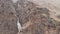 An aerial view of a young photographer with a DSLR camera in his hand stands high on a rock against a waterfall in a