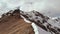 Aerial view of a young thin man with a backpack and trekking sticks climbs uphill on a crest on the border of rocks and