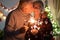 An affectionate kiss between elderly wife and husband who celebrate Christmas with sparks. Lights and Christmas tree in the