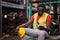 Africa American holding safety helmet and sitting at forklift in the automotive part warehouse. Worker look at smartphone and get