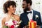 African american bride holding fowers near happy bridegroom with present