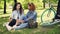 African American girl is chatting with her Caucasian friend sitting on grass in park, friends are talking and smiling
