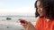 African American girl teenager young woman wearing orange hoodie, sitting on a sea front using her smart cell phone