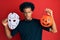 African american man with afro hair wearing hockey mask and halloween pumpking in shock face, looking skeptical and sarcastic,