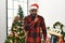 African american man wearing santa claus hat standing by christmas tree hand on mouth telling secret rumor, whispering malicious