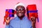 African American man wearing stylish plaid shirt great smile in santa hat with gift box on pink background studio.dark