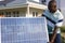 African american mid adult man laughing and carrying solar panel against house in yard on sunny day