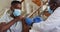 African american senior male doctor giving covid vaccine to male patient in home, wearing face masks