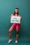 African american sportswoman smiling, looking at camera and presenting placard with love your body inscription on green