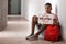 African-American teenage boy holding sheet of paper with word BULLYING while sitting on floor at school