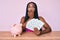 African american woman with braids sitting on the table with piggy bank and dollars covering mouth with hand, shocked and afraid