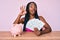 African american woman with braids sitting on the table with piggy bank and dollars doing ok sign with fingers, smiling friendly