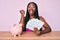 African american woman with braids sitting on the table with piggy bank and dollars screaming proud, celebrating victory and