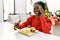 African american woman eating hamburger talking on smartphone sitting on table by christmas tree at home