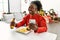 African american woman eating hamburger using smartphone sitting on table by christmas tree at home